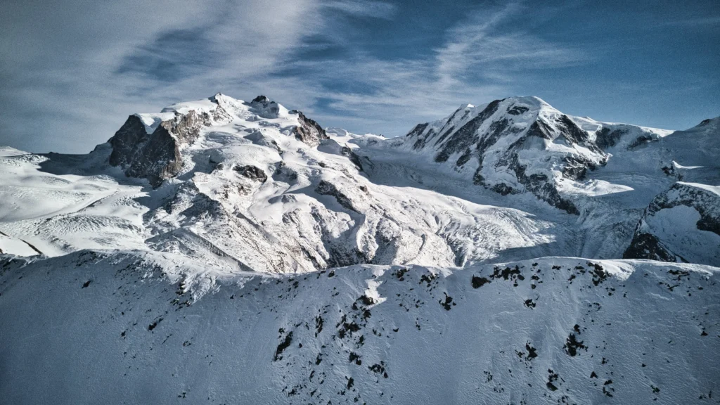 Schneebedecktes Monte Rosa-Gebirgsmassiv teilweise mit Wolken und blauem Himmel im Hintergrund. Foto von Daniel R.-Unsplash