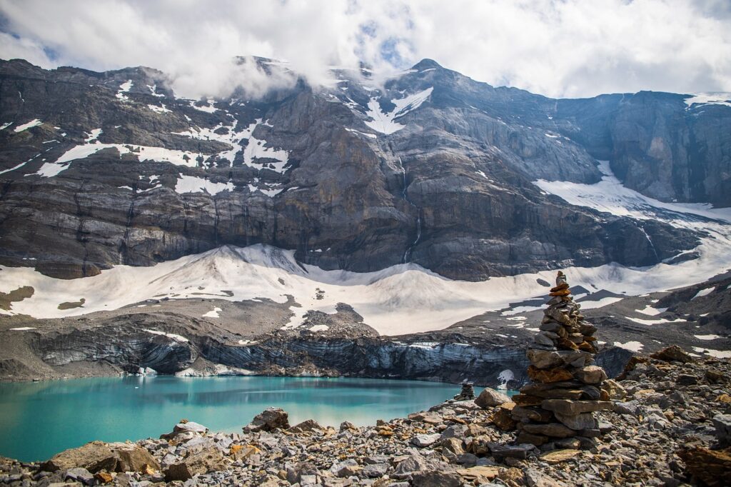 Auf dem Foto sind hohe Berge zu sehen. Sie sind zum Teil mit Schnee bedeckt. Unten links auf dem Bild ist ein See zu sehen. Das Wasser im See hat eine wunderschöne grüne Farbe. Der Himmel ist von Wolken bedeckt. 