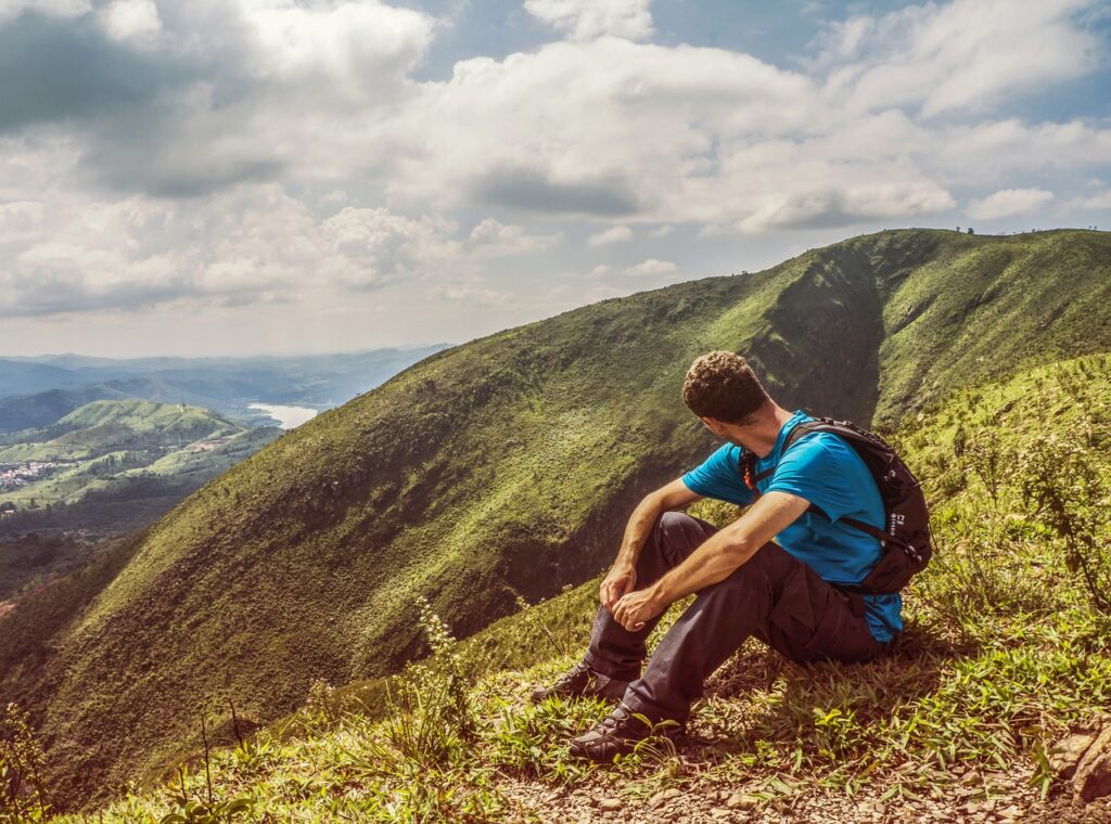 Auf dem Foto ist ein junger Mann zu sehen. Er wandern durch den Alpe Adria Trail und macht grade eine Wanderpause auf dem Gipfle eines Berges und bewundert die Natur. 